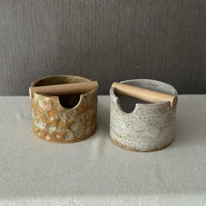 Op de afbeelding: Twee cilindrische keramische houders met een inkeping in de rand en een horizontale houten handgreep aan de bovenzijde. De ene houder heeft een textuur met een bruin en beige glazuur, de andere heeft een gespikkelde lichtgrijze en witte afwerking.