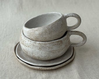 May include: Two speckled ceramic coffee cups with handles are stacked on a matching saucer. The cups and saucer are off-white with brown speckles, and the cups have a rounded shape.