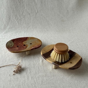 Può includere: Due portasapone ovali in ceramica con piccoli piedini e fori di drenaggio. Uno presenta una smaltatura marmorizzata rossa e marrone chiaro, l'altro ha strisce marroni e beige e sostiene una spazzola rotonda in legno con setole naturali chiare.