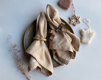 May include: Two beige linen napkins with a wooden napkin ring on a brown plate. The napkins are folded in a decorative way and tied with a green and red ribbon. There are three wooden ornaments on a white background.