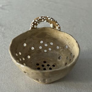 May include: A small, beige ceramic colander with a handle. The colander has numerous small holes and a handle decorated with white, textured accents. The bowl is round with a slightly irregular rim.