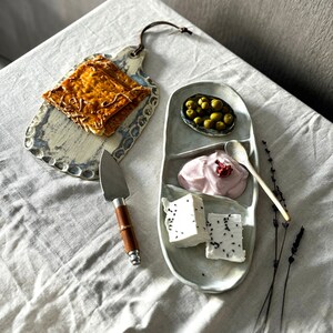 May include: A white ceramic serving platter with three sections. The sections contain a small bowl of green olives, a dollop of pink yogurt, and two pieces of white cheese with black sesame seeds. A wooden handled butter knife and a small wooden spoon are also included.