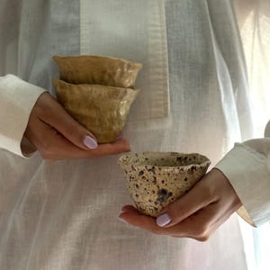 K&ouml;nnte beinhalten: Drei handgefertigte Keramikschalen in Beige- und Braunt&ouml;nen. Zwei Schalen sind gestapelt, die dritte wird separat gehalten. Die Schalen haben eine strukturierte, organische Form, mit einem gesprenkelten Muster auf der dritten. Die Schalen werden vor einem wei&szlig;en Hintergrund gehalten.