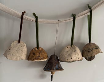 May include: Five ceramic bell ornaments hanging from a cream-colored fabric strip. Two are speckled off-white, one is textured brown, one is dark blue with a small clapper, and one is a mushroom-shaped bell with a dark line detail and green ribbon.