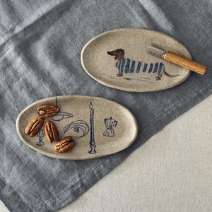 May include: Two oval ceramic serving dishes with hand-painted designs. One features a dachshund in blue stripes, the other has a candle, coffee pot, and other illustrations. Pecans and a fork with a wooden handle are also present.