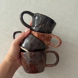 May include: A stack of four handmade ceramic mugs in varying shades of brown and black. Each mug has a unique textured surface and a curved handle. The mugs are stacked together, showcasing their different colors and designs.