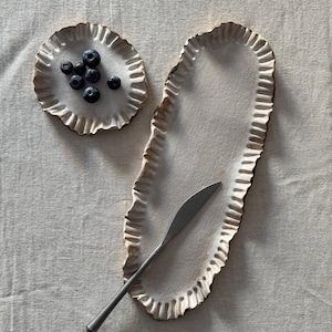 May include: Two white ceramic serving dishes with ruffled edges. The smaller dish has blueberries on it. The larger dish has a silver knife on it.