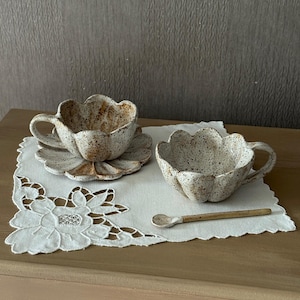 K&ouml;nnte beinhalten: Zwei gesprenkelte Keramik-Tassen und Untertassen mit floralem Design, begleitet von einem kleinen L&ouml;ffel. Die Tassen und Untertassen sind wei&szlig; mit braunen Sprenkeln und einem bl&uuml;tenblattartigen Rand. Sie liegen auf einer wei&szlig;en, bestickten Serviette.