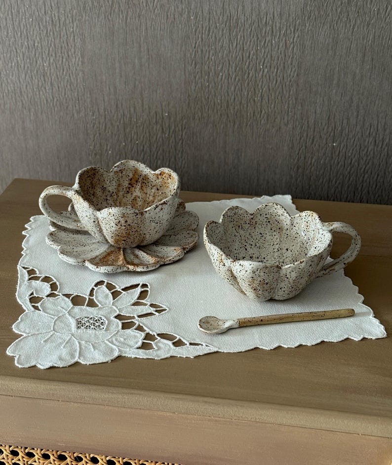 K&ouml;nnte beinhalten: Zwei gesprenkelte Keramik-Tassen und Untertassen mit floralem Design, begleitet von einem kleinen L&ouml;ffel. Die Tassen und Untertassen sind wei&szlig; mit braunen Sprenkeln und einem bl&uuml;tenblattartigen Rand. Sie liegen auf einer wei&szlig;en, bestickten Serviette.