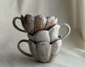 Può includere: Tre tazze in ceramica impilate con una glassa bianco sporco macchiettata e un design a bordo a forma di petalo. Ogni tazza ha un manico curvo e un aspetto rustico fatto a mano. Le tazze sono disposte su uno sfondo di tessuto dai toni neutri.