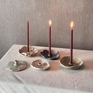 Könnte beinhalten: Drei Kerzenhalter aus Keramik mit drei roten Kerzen. Die Kerzenhalter haben unterschiedliche Formen und Größen und sind alle aus rauer, strukturierter Keramik gefertigt.