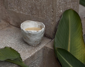 May include: A small, hand-formed ceramic cup with an irregular, organic rim and a textured white glaze featuring brown, earthy accents. The cup has a slightly flared shape and a visible base, containing a light brown liquid inside the vessel.