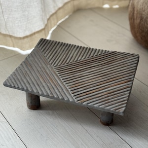 Könnte beinhalten: Ein graues dekoratives Holztablett mit rechteckiger Oberseite und drei kurzen, zylindrischen Beinen. Die Oberfläche des Tabletts weist ein strukturiertes, geometrisches Design mit diagonalen und parallelen Linien auf. Das Tablett ist aus Holz und hat ein rustikales Aussehen.