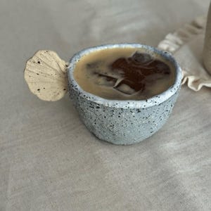 Puede incluir: Una taza de café de cerámica gris moteada con un asa única en forma de hoja. La taza está llena de café y hielo. La taza está colocada sobre una superficie de color neutro, lo que sugiere una estética minimalista. El diseño de la taza es artesanal.
