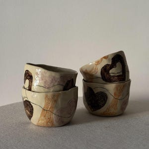 May include: Three small, off-white ceramic bowls with irregular, hand-formed edges. The bowls feature hand-drawn illustrations in dark brown, tan, and muted purple, including heart shapes and abstract lines, arranged in a stacked and grouped formation.