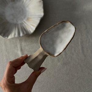 Puede incluir: Una cuchara de cerámica hecha a mano con interior blanco y exterior beige. La cuchara tiene un cuenco ancho y poco profundo y un mango corto y texturizado. Un cuenco de cerámica a juego con un borde con volantes está en el fondo.