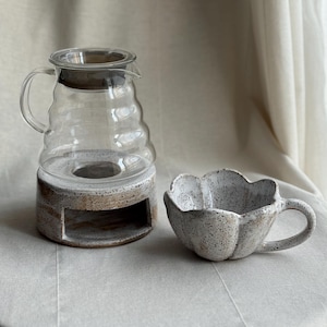 K&ouml;nnte beinhalten: Eine Kaffeekaraffe aus klarem Glas mit dunklem Deckel steht auf einem gesprenkelten Keramiksockel. Daneben steht eine passende Keramiktasse mit bl&uuml;tenblattartigem Design und Henkel. Das Set steht auf einer neutralfarbenen Oberfl&auml;che.