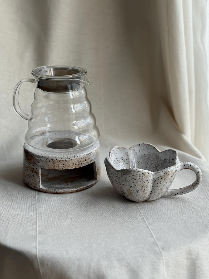 K&ouml;nnte beinhalten: Eine Kaffeekaraffe aus klarem Glas mit dunklem Deckel steht auf einem gesprenkelten Keramiksockel. Daneben steht eine passende Keramiktasse mit bl&uuml;tenblattartigem Design und Henkel. Das Set steht auf einer neutralfarbenen Oberfl&auml;che.
