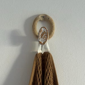 Puede incluir: Un anillo de toalla de cerámica con un acento ovalado decorativo con un diseño de árbol. El anillo está unido a una toalla marrón en forma de red. El anillo y la toalla cuelgan de una pared blanca.