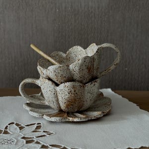 K&ouml;nnte beinhalten: Zwei gestapelte, gesprenkelte Keramik-Teetassen mit passenden Untertassen. Die Tassen und Untertassen sind wie Bl&uuml;tenbl&auml;tter geformt. Ein kleiner Holzl&ouml;ffel liegt in der oberen Tasse. Die Teetassen und Untertassen sind wei&szlig; mit braunen Sprenkeln.