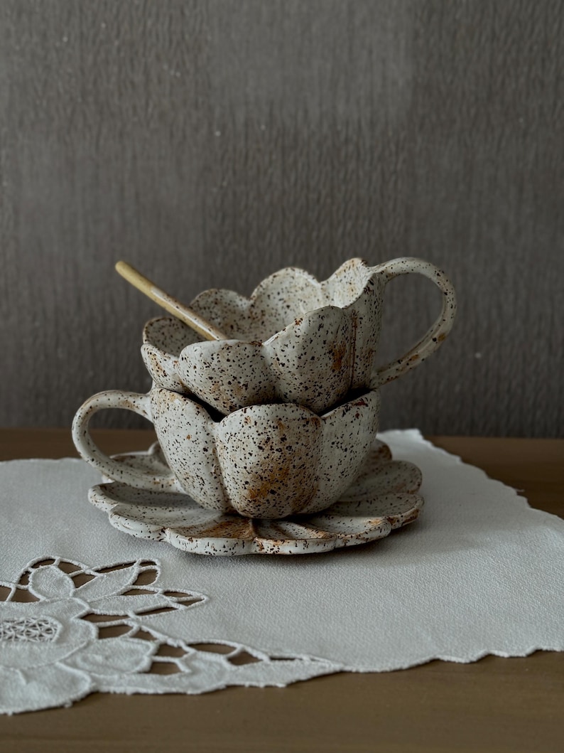 K&ouml;nnte beinhalten: Zwei gestapelte, gesprenkelte Keramik-Teetassen mit passenden Untertassen. Die Tassen und Untertassen sind wie Bl&uuml;tenbl&auml;tter geformt. Ein kleiner Holzl&ouml;ffel liegt in der oberen Tasse. Die Teetassen und Untertassen sind wei&szlig; mit braunen Sprenkeln.