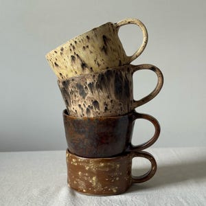 May include: A vertical stack of four ceramic mugs with handles. The top mug is cream with dark brown speckles, the second is dark brown with a drip glaze, the third is a solid dark brown, and the bottom mug is a light brown with yellow speckles.