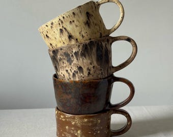 May include: A vertical stack of four ceramic mugs with handles. The top mug is cream with dark brown speckles, the second is dark brown with a drip glaze, the third is a solid dark brown, and the bottom mug is a light brown with yellow speckles.