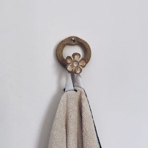 Könnte beinhalten: Ein Keramikhaken in Blumenform mit brauner Glasur hängt an einer weißen Wand. Ein beigefarbenes Handtuch mit schwarzem Rand hängt an dem Haken.