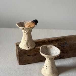 May include: Two off-white ceramic incense holders with a speckled finish and flared rims. Each holder features a textured, tapered base. One holder contains a piece of charred wood, while the other is empty, showcasing the wide, shallow bowl design.