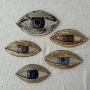 Puede incluir: Cinco piezas decorativas de cerámica con forma de ojo. Cada pieza tiene una paleta de colores única, con variaciones de marrón, gris y blanco. Los iris son azul oscuro, marrón y negro. Las piezas están dispuestas sobre una superficie de color neutro.