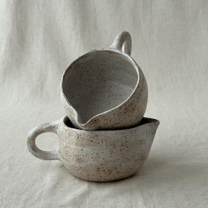 Può includere: Due brocche in ceramica maculata, una sovrapposta all'altra. Le brocche hanno una forma arrotondata con un beccuccio e un manico curvo. La ceramica è di colore bianco sporco naturale con macchie marroni.