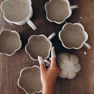 K&ouml;nnte beinhalten: Eine Sammlung handgefertigter, wolkenf&ouml;rmiger Keramikbecher und eine passende Untertasse in einem neutralen Beige. Die Becher haben einen Henkel und sind auf einer Holzoberfl&auml;che angeordnet. Das Bild zeigt das einzigartige Design und die Textur der Keramik.