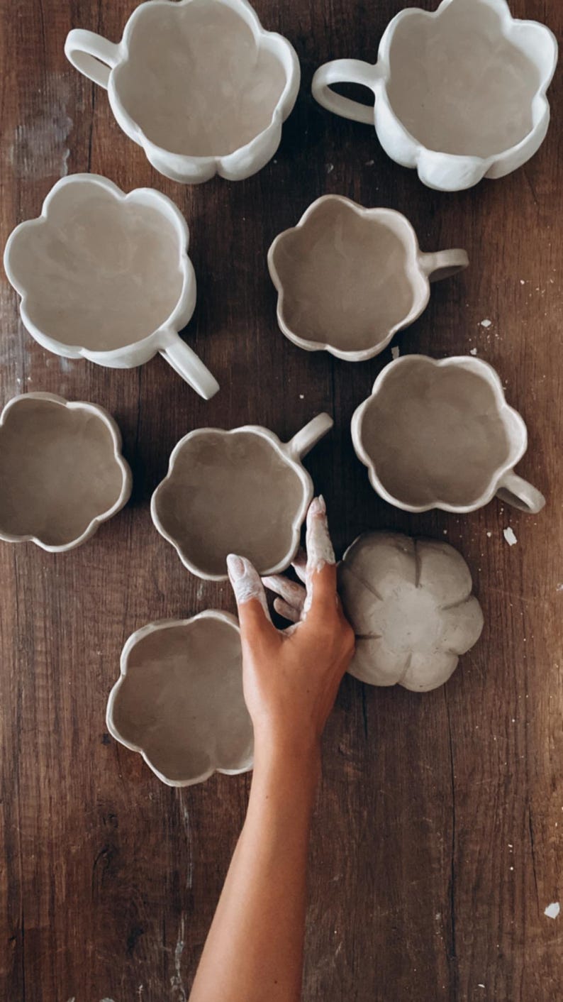 K&ouml;nnte beinhalten: Eine Sammlung handgefertigter, wolkenf&ouml;rmiger Keramikbecher und eine passende Untertasse in einem neutralen Beige. Die Becher haben einen Henkel und sind auf einer Holzoberfl&auml;che angeordnet. Das Bild zeigt das einzigartige Design und die Textur der Keramik.