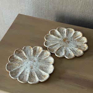 K&ouml;nnte beinhalten: Zwei wei&szlig;e Keramikuntersetzer in Form einer G&auml;nseblume. Die Untersetzer haben eine gesprenkelte, strukturierte Oberfl&auml;che mit braunen und beigen Akzenten. Sie liegen auf einer braunen Holzoberfl&auml;che.