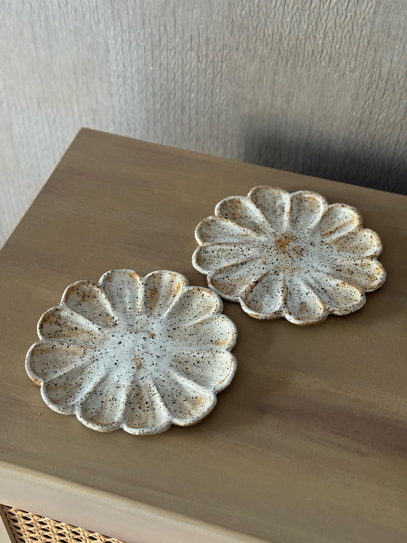 K&ouml;nnte beinhalten: Zwei wei&szlig;e Keramikuntersetzer in Form einer G&auml;nseblume. Die Untersetzer haben eine gesprenkelte, strukturierte Oberfl&auml;che mit braunen und beigen Akzenten. Sie liegen auf einer braunen Holzoberfl&auml;che.