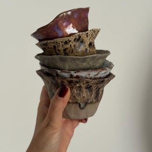 K&ouml;nnte beinhalten: Ein Stapel handgefertigter Keramikschalen in verschiedenen Erdt&ouml;nen. Die oberste Schale ist ein tiefes Burgunderrot mit gl&auml;nzendem Finish, w&auml;hrend die anderen gesprenkelte und strukturierte Designs aufweisen. Die Schalen werden in einer Hand gehalten.