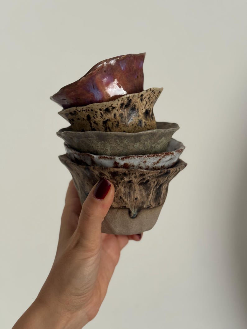 K&ouml;nnte beinhalten: Ein Stapel handgefertigter Keramikschalen in verschiedenen Erdt&ouml;nen. Die oberste Schale ist ein tiefes Burgunderrot mit gl&auml;nzendem Finish, w&auml;hrend die anderen gesprenkelte und strukturierte Designs aufweisen. Die Schalen werden in einer Hand gehalten.