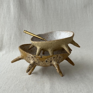 以下が含まれることがあります： ベージュと茶色の斑点模様がある陶器のボウル2つが重なっており、それぞれ側面に4本の尖った脚が付いています。ボウルの内側は白地に暗い斑点があります。上のボウルの中に、細い金色の金属製スプーンが入っています。