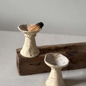 May include: Two off-white ceramic incense holders with a speckled finish and a flared, uneven rim. Each holder features a narrow neck with a textured band. One holder contains a piece of charred wood, while the other is empty.