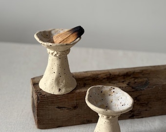May include: Two off-white ceramic incense holders with a speckled finish and a flared, uneven rim. Each holder features a narrow neck with a textured band. One holder contains a piece of charred wood, while the other is empty.