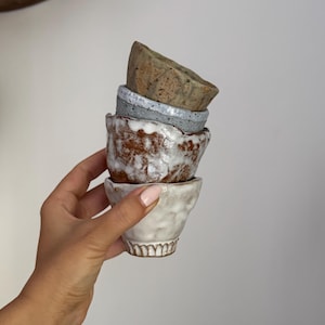 Può includere: Una pila di quattro piccole ciotole in ceramica fatte a mano, in vari colori e trame. Le ciotole sono impilate l'una sull'altra, tenute da una mano. I colori includono bianco, marrone, grigio e beige maculato. Le ciotole hanno un'estetica rustica e organica.