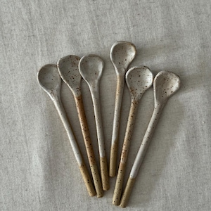 K&ouml;nnte beinhalten: Eine Sammlung von sechs handgefertigten Keramik-L&ouml;ffeln. Jeder L&ouml;ffel hat eine gesprenkelte, cremefarbene Schale und einen Griff mit einem gesprenkelten, cremefarbenen oberen Teil und einem beigefarbenen unteren Teil. Die L&ouml;ffel sind auf einer neutralfarbenen Leinenoberfl&auml;che angeordnet.