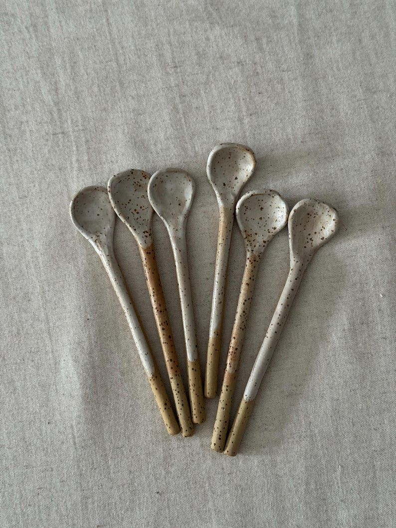 K&ouml;nnte beinhalten: Eine Sammlung von sechs handgefertigten Keramik-L&ouml;ffeln. Jeder L&ouml;ffel hat eine gesprenkelte, cremefarbene Schale und einen Griff mit einem gesprenkelten, cremefarbenen oberen Teil und einem beigefarbenen unteren Teil. Die L&ouml;ffel sind auf einer neutralfarbenen Leinenoberfl&auml;che angeordnet.
