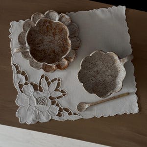 K&ouml;nnte beinhalten: Ein Keramik-Tee-Tassen- und Untertassen-Set mit gesprenkelter, cremefarbener Oberfl&auml;che, platziert auf einer wei&szlig;en, bestickten Tischdecke. Ein kleiner passender L&ouml;ffel liegt neben der Tasse. Das Set steht auf einer Holzoberfl&auml;che.