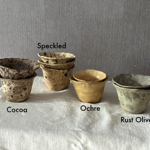 K&ouml;nnte beinhalten: Eine Sammlung handgefertigter Keramikbecher in verschiedenen Farben und Texturen. Die Becher sind mit "Cocoa", "Speckled", "Ochre" und "Rust Olive" beschriftet. Jeder Becher hat eine einzigartige gesprenkelte oder strukturierte Oberfl&auml;che.