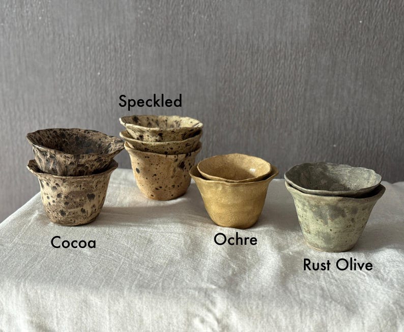 K&ouml;nnte beinhalten: Eine Sammlung handgefertigter Keramikbecher in verschiedenen Farben und Texturen. Die Becher sind mit "Cocoa", "Speckled", "Ochre" und "Rust Olive" beschriftet. Jeder Becher hat eine einzigartige gesprenkelte oder strukturierte Oberfl&auml;che.