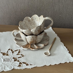 K&ouml;nnte beinhalten: Ein Keramik-Tassen- und Untertassen-Set mit gesprenkelter, cremefarbener Oberfl&auml;che. Die Tasse und die Untertasse haben einen gewellten Rand, und ein kleiner passender L&ouml;ffel liegt daneben. Das Set steht auf einer wei&szlig;en, bestickten Serviette.