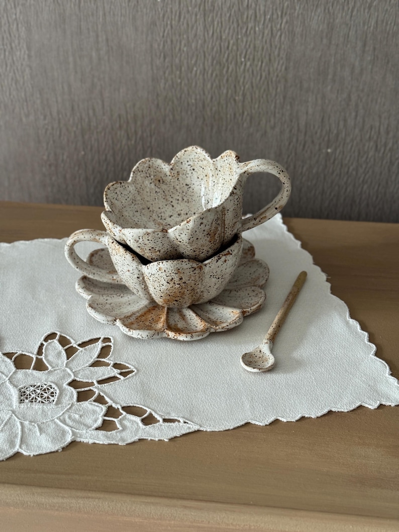 K&ouml;nnte beinhalten: Ein Keramik-Tassen- und Untertassen-Set mit gesprenkelter, cremefarbener Oberfl&auml;che. Die Tasse und die Untertasse haben einen gewellten Rand, und ein kleiner passender L&ouml;ffel liegt daneben. Das Set steht auf einer wei&szlig;en, bestickten Serviette.