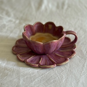 Könnte beinhalten: Ein Keramik-Tassen- und Untertassen-Set in Form einer Blume mit abgerundeten Blütenblättern. Das Geschirr hat eine glänzende, violett-magenta farbene Glasur mit cremefarbenen Rändern. Die Tasse enthält eine hellbraune Flüssigkeit und hat einen Henkel.