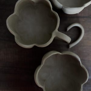 K&ouml;nnte beinhalten: Drei handgefertigte, unglasierte Keramikst&uuml;cke in Blumenform. Eines ist eine Tasse mit gebogenem Henkel, die anderen beiden sind Schalen. Die St&uuml;cke haben einen neutralen, erdigen Farbton, vor dunklem Holzgrund.